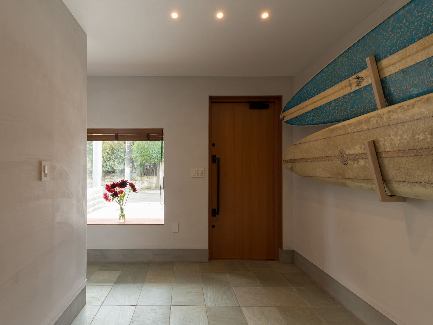 玄関扉の横に花が飾られた出窓がある、壁にサーフボードが飾られた玄関・デザイン住宅 注文住宅・デザイン住宅建築実例_『CASE643 だて窓のハウス』_玄関